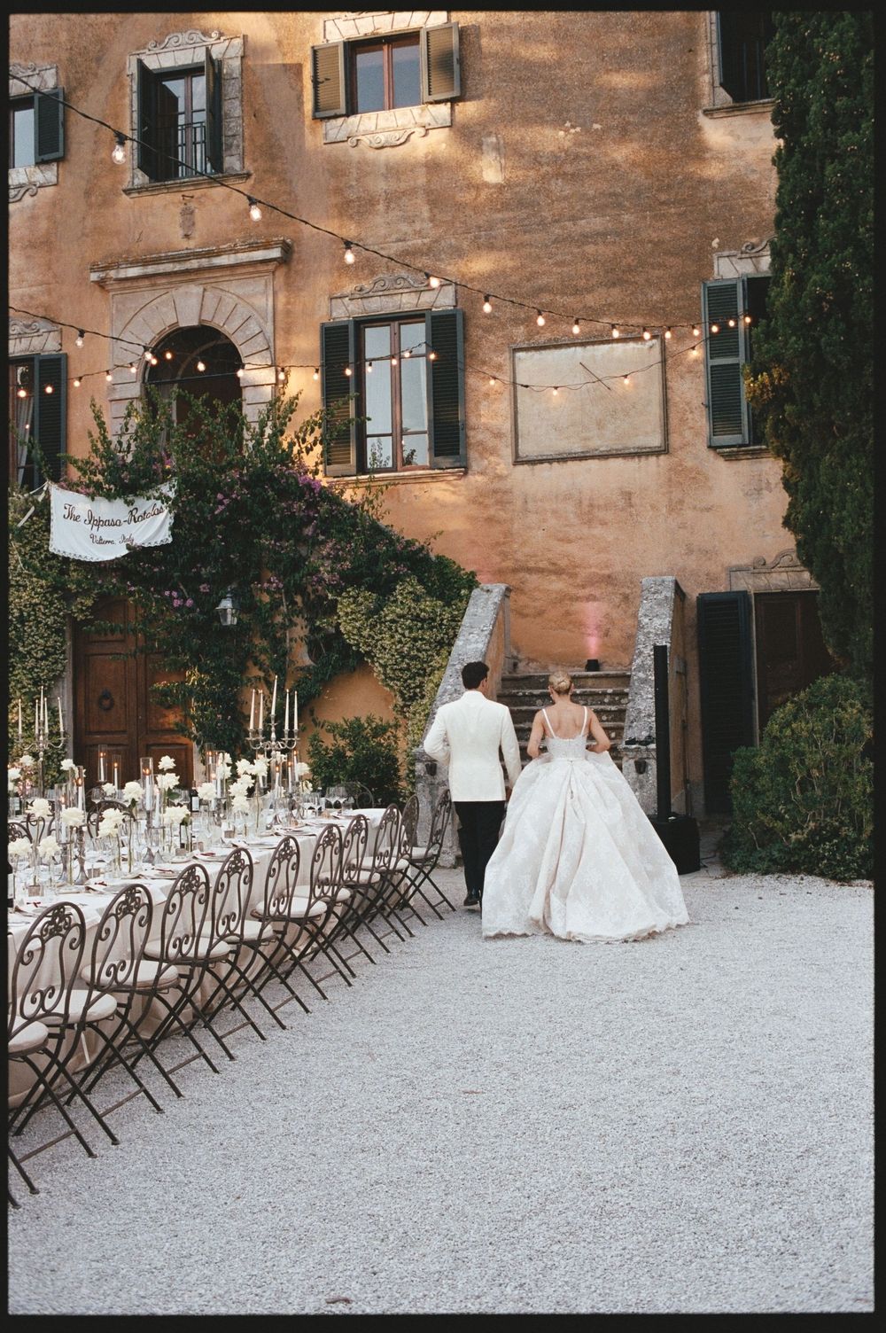 Bride and groom walk toward rustic Italian villa with elegant outdoor reception table setting at twilight.