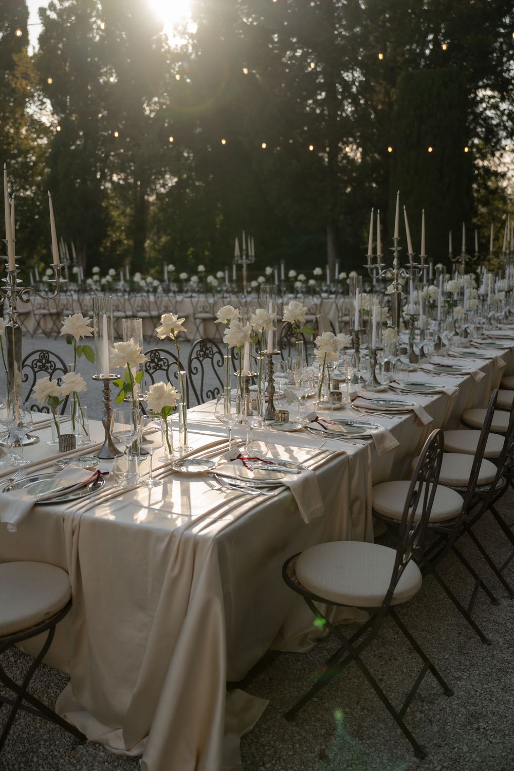 Elegant outdoor wedding reception with long tables, white linens, candelabras, and floral centerpieces bathed in golden sunlight.
