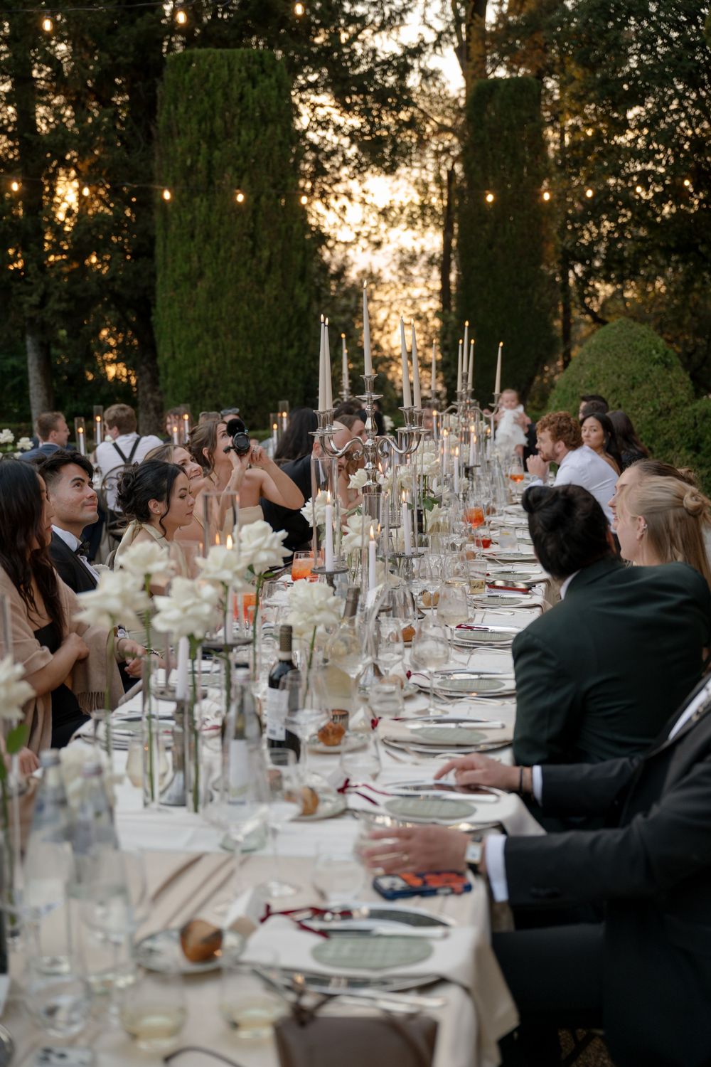 Elegant outdoor wedding reception dinner with long table, candles, and floral arrangements at sunset.