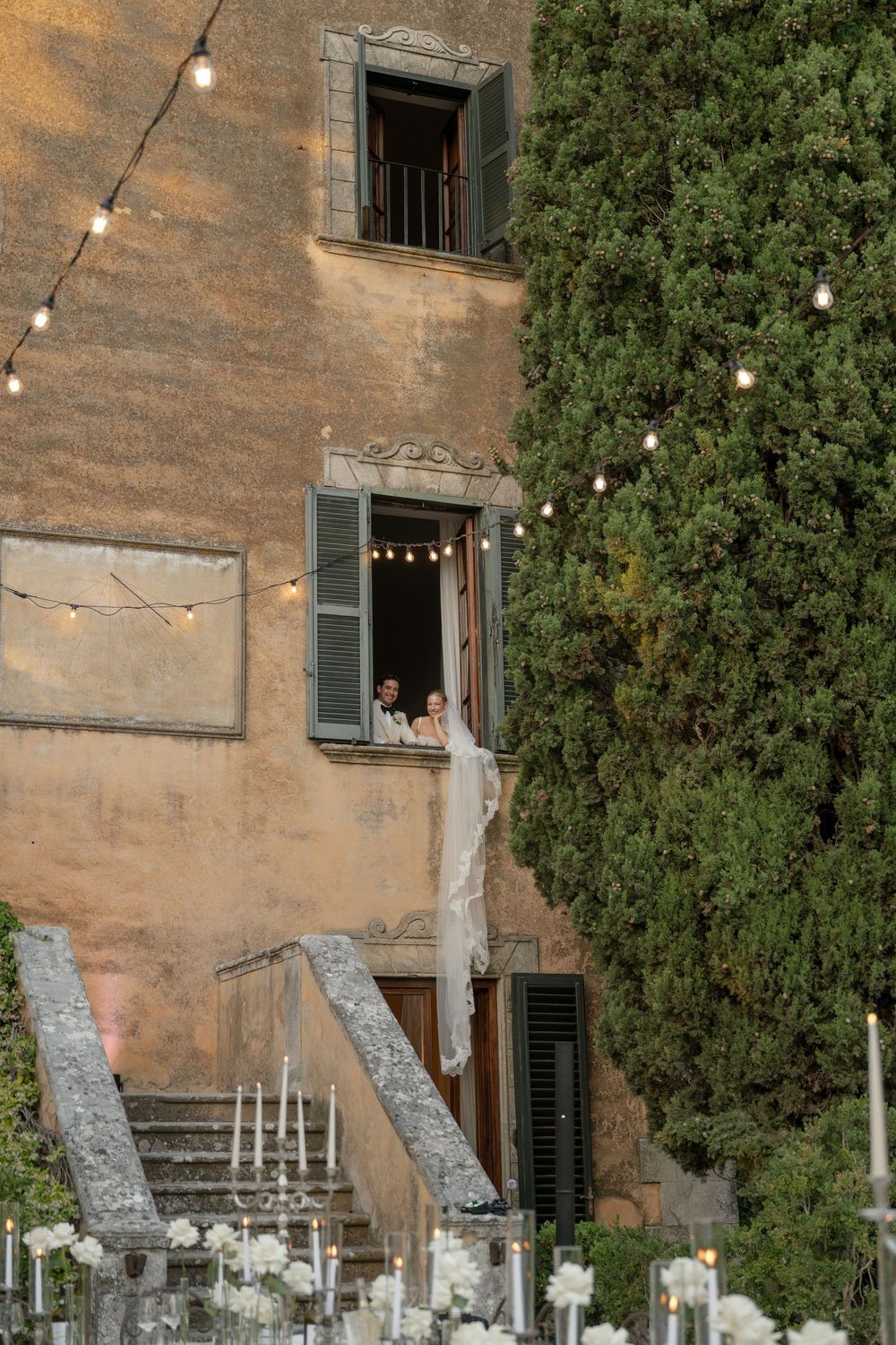 Bride and groom appear at window of elegant Italian villa adorned with string lights at dusk.