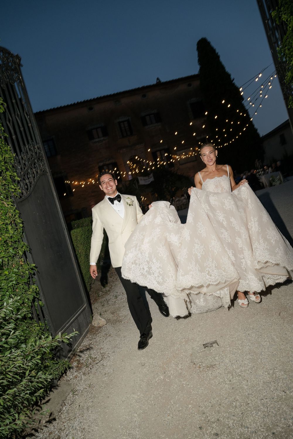 Bride and groom walk through iron gates at dusk, groom carrying wedding dress train, string lights glowing behind them.