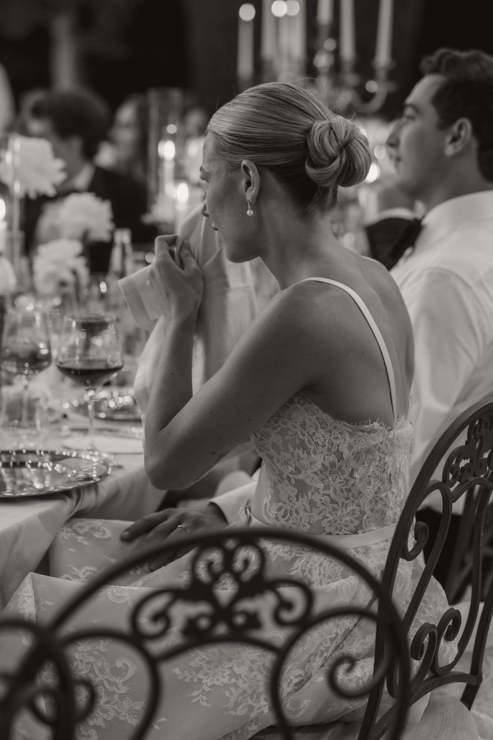 Black and white photo of bride in lace dress seated at wedding reception table, listening attentively to speeches.