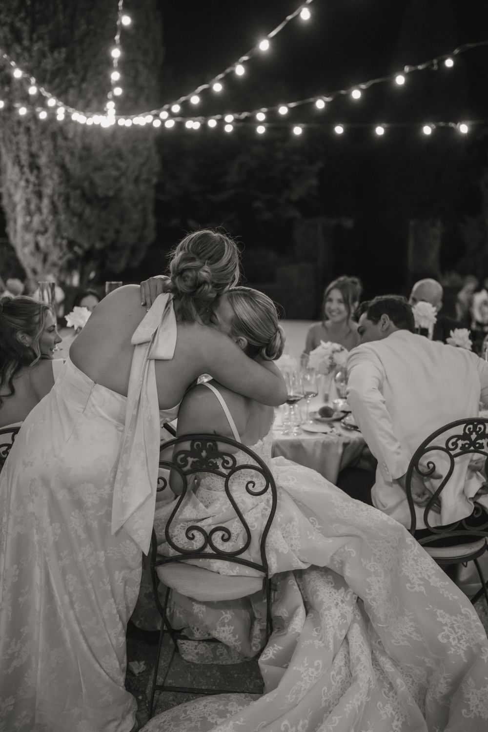 Two brides share an emotional embrace at an outdoor wedding reception under string lights in a black and white photo.