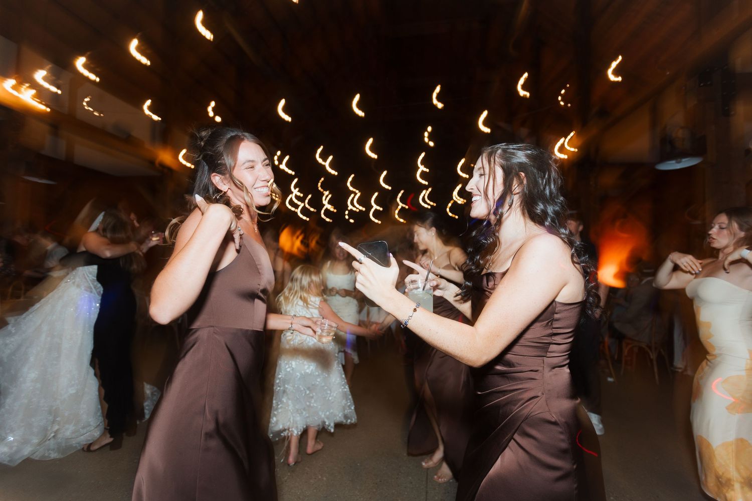 Two bridesmaids in brown satin dresses dancing energetically at a wedding reception with warm lights.