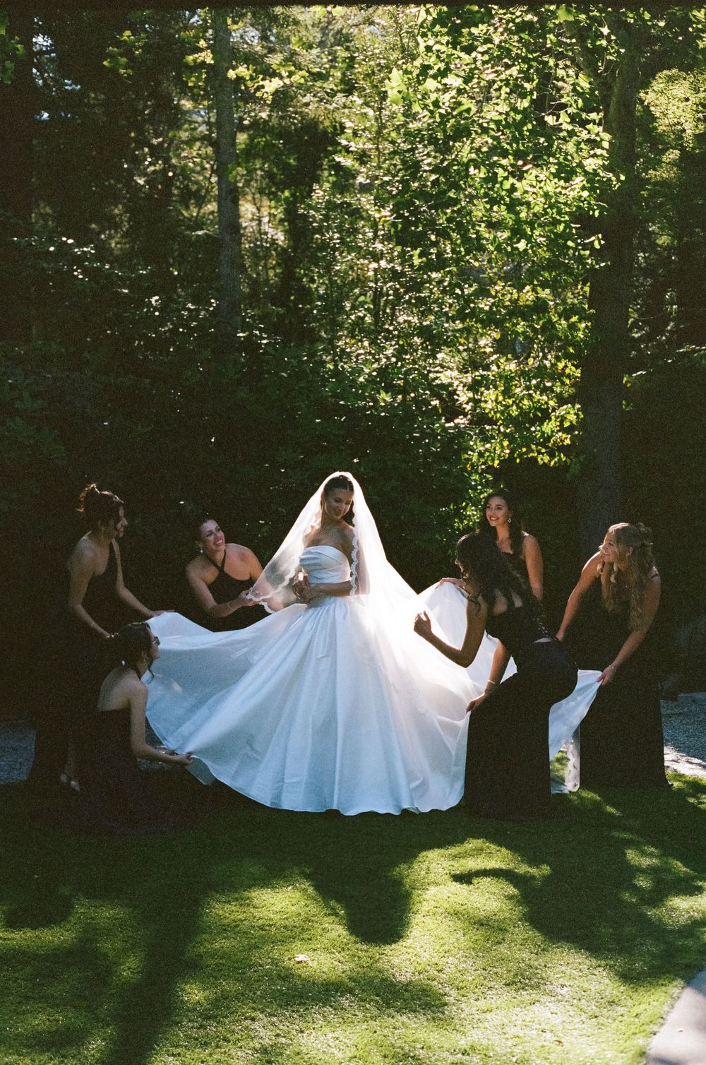 Bridesmaids in black dresses fluff the bride's white ball gown in a sunlit garden setting.