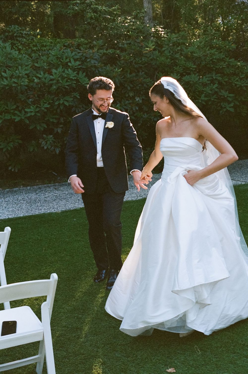 Bride in strapless white ball gown and groom in tuxedo walk hand in hand on green lawn at outdoor wedding.