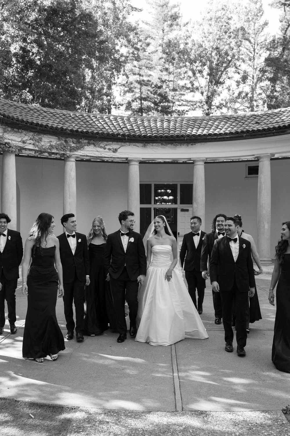 Bride and groom walk with wedding party in black and white photo near elegant columned rotunda.