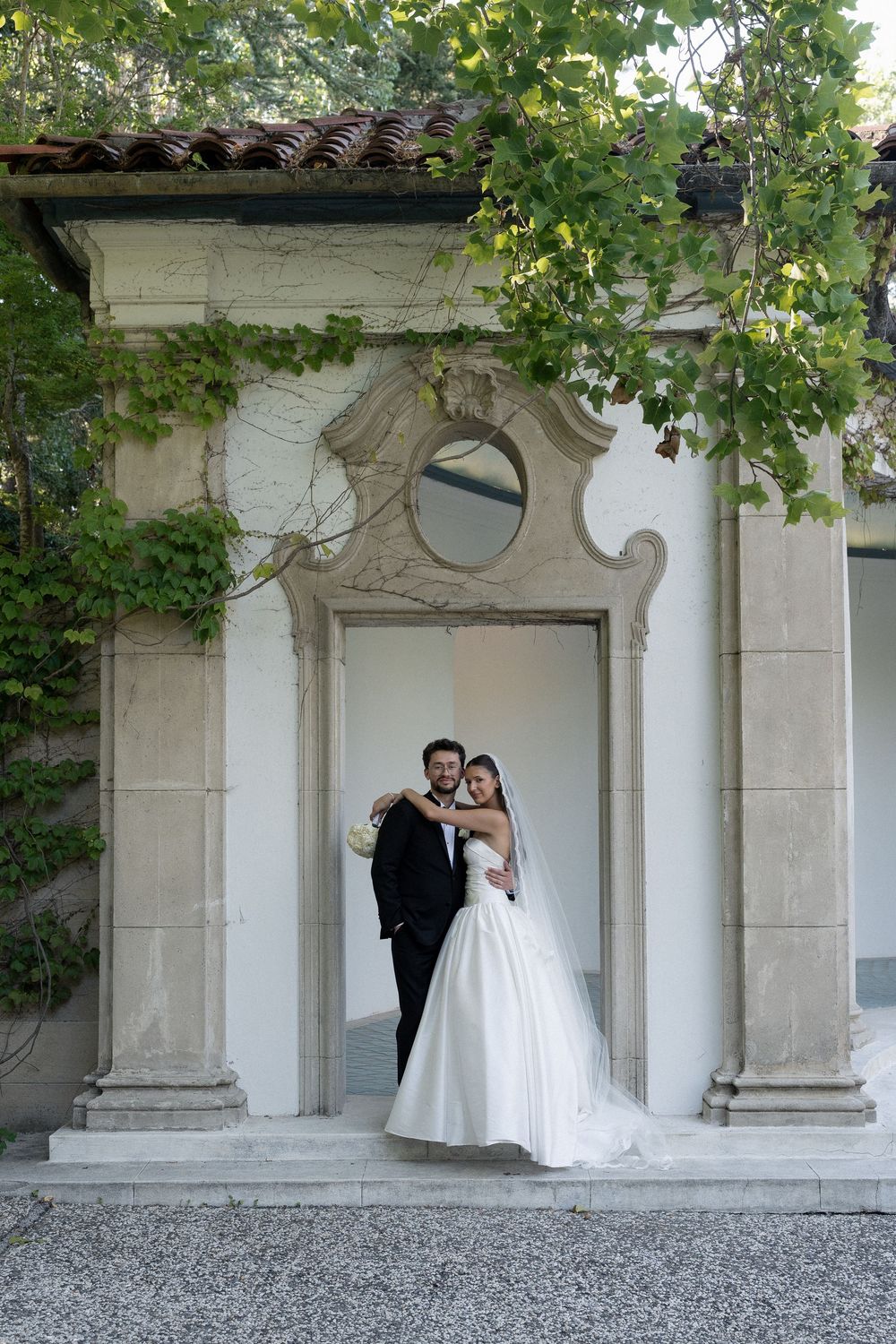 Newlyweds embrace in a white stone archway surrounded by lush greenery at their wedding venue.