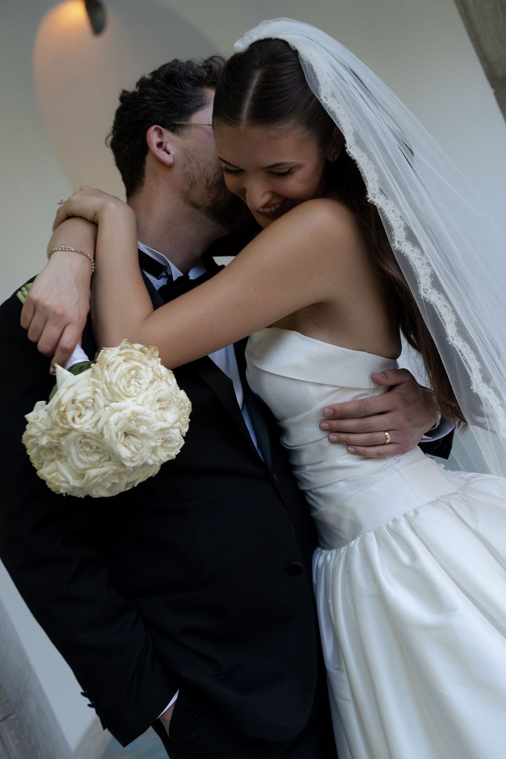 Newlywed bride and groom sharing tender embrace with white rose bouquet after romantic wedding ceremony.