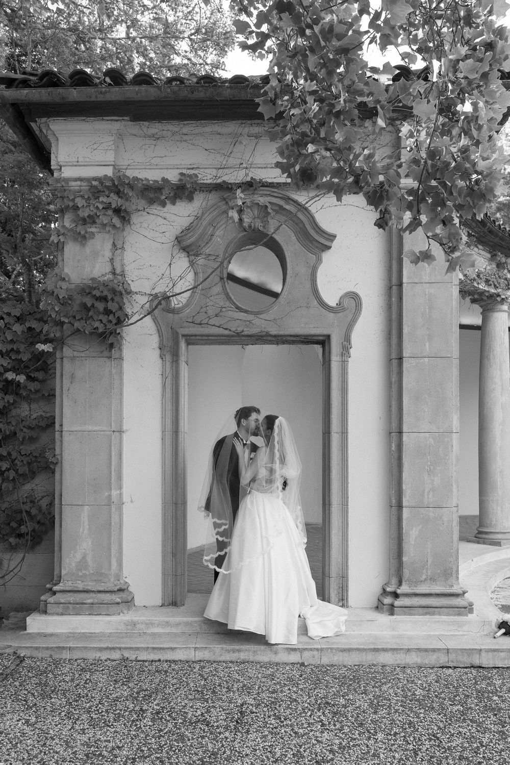 Black and white photo of bride and groom kissing in an ornate stone doorway surrounded by ivy.
