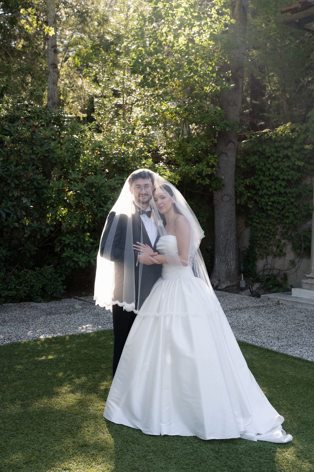 Bride and groom embrace under flowing veil in sunlit garden with lush green trees.