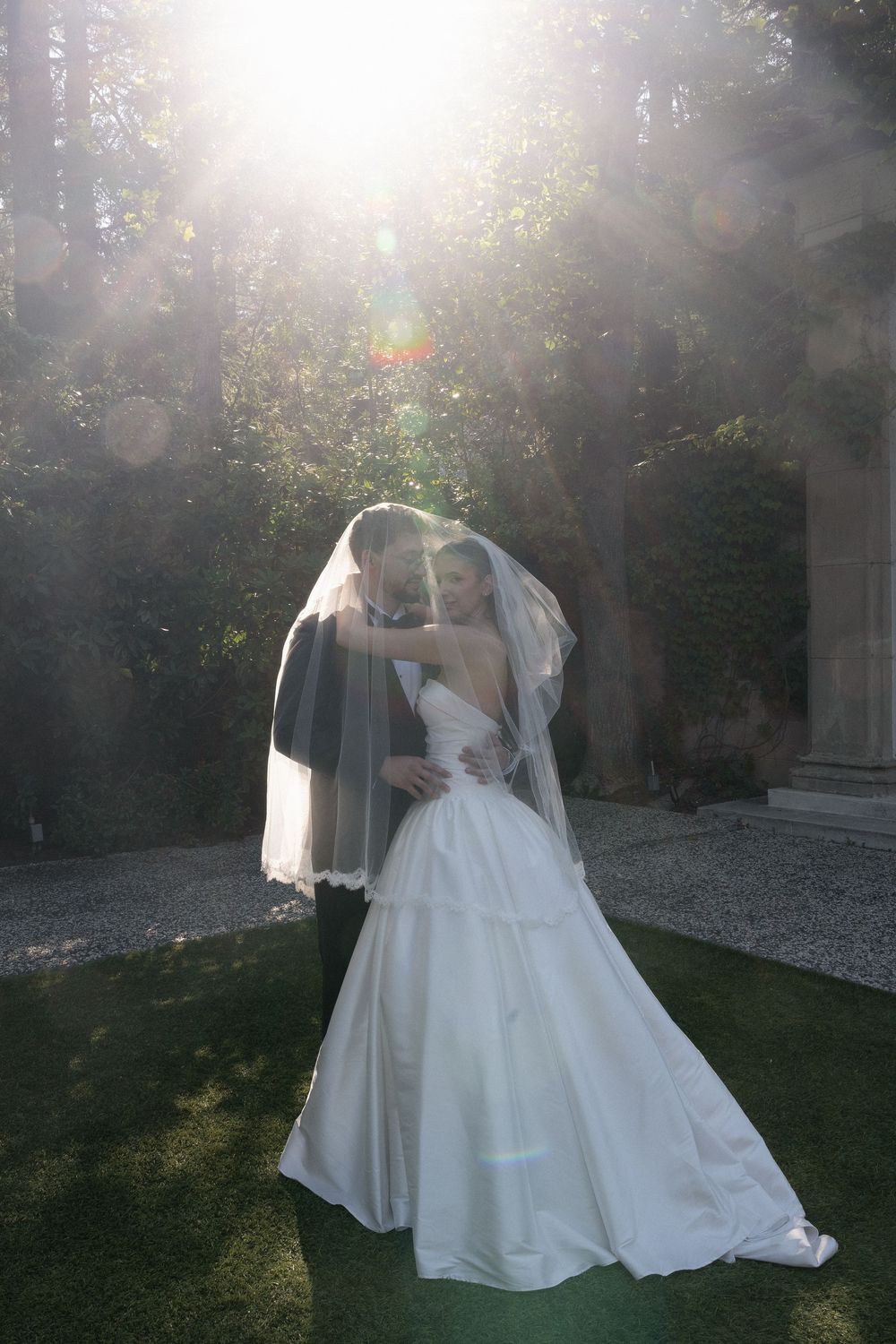 Couple shares romantic kiss under bridal veil with beautiful sunlight flare in garden.