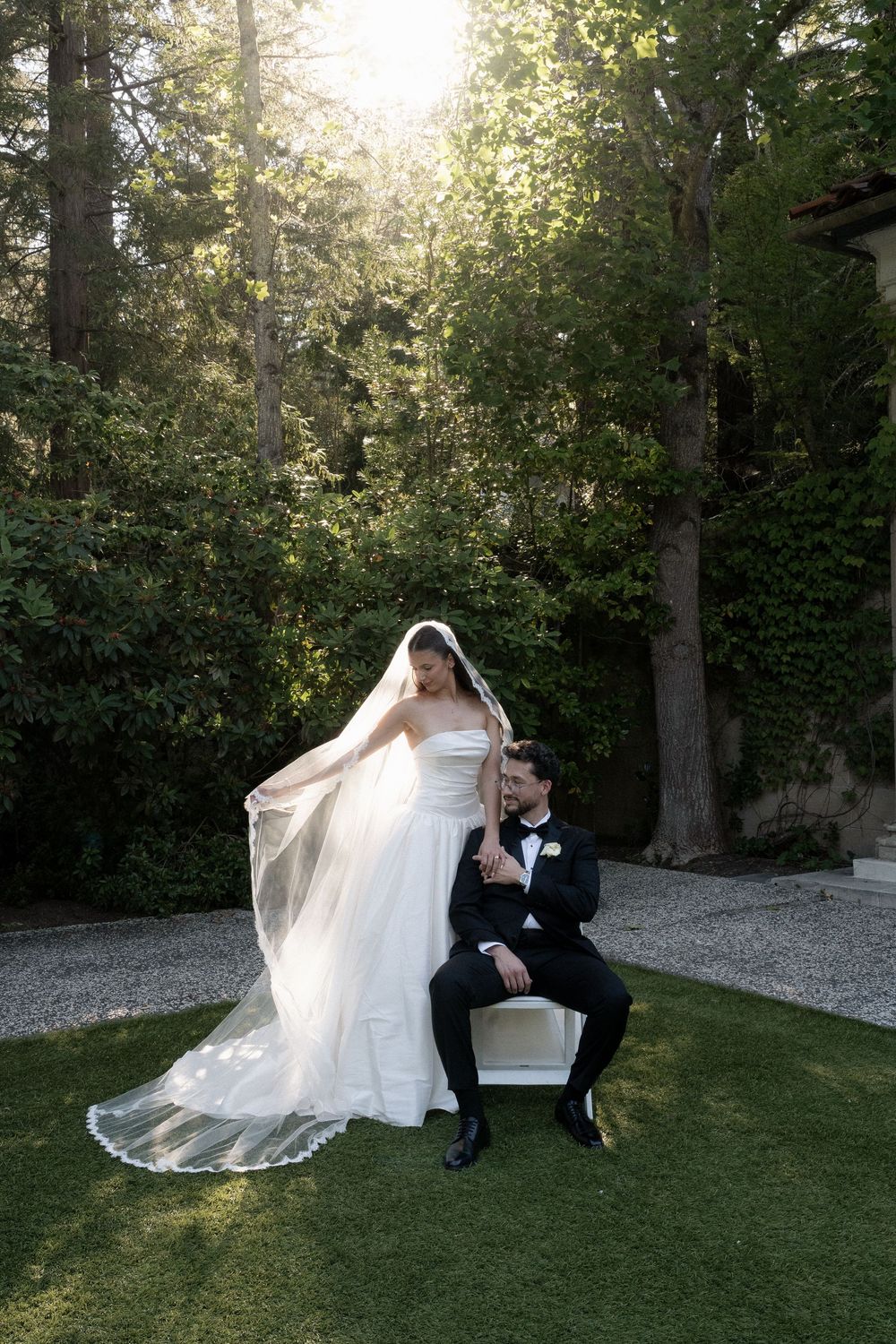 Bride in white gown and veil stands beside seated groom in black tuxedo in a sunlit garden.