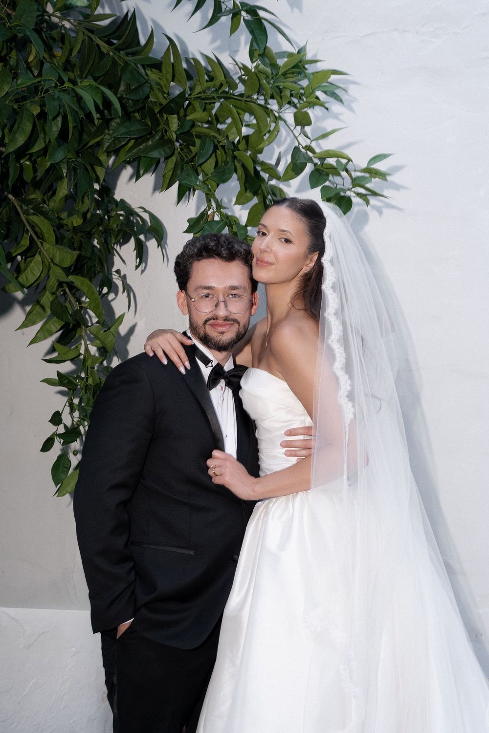 A bride and groom pose together under green foliage, she wears a white gown and veil, he wears a black tuxedo.