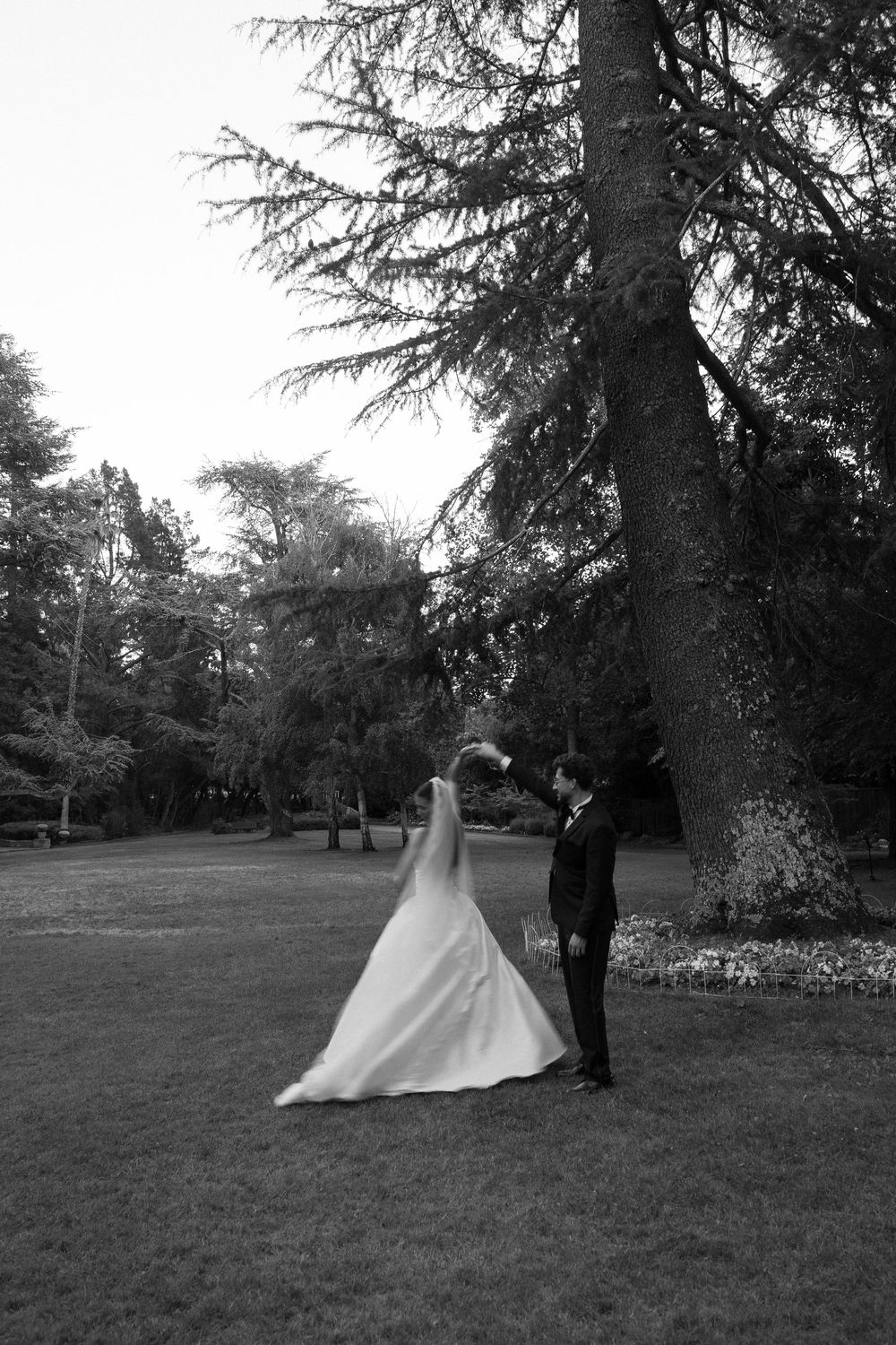 Black and white photo of bride and groom dancing outdoors near large trees in a lush garden.