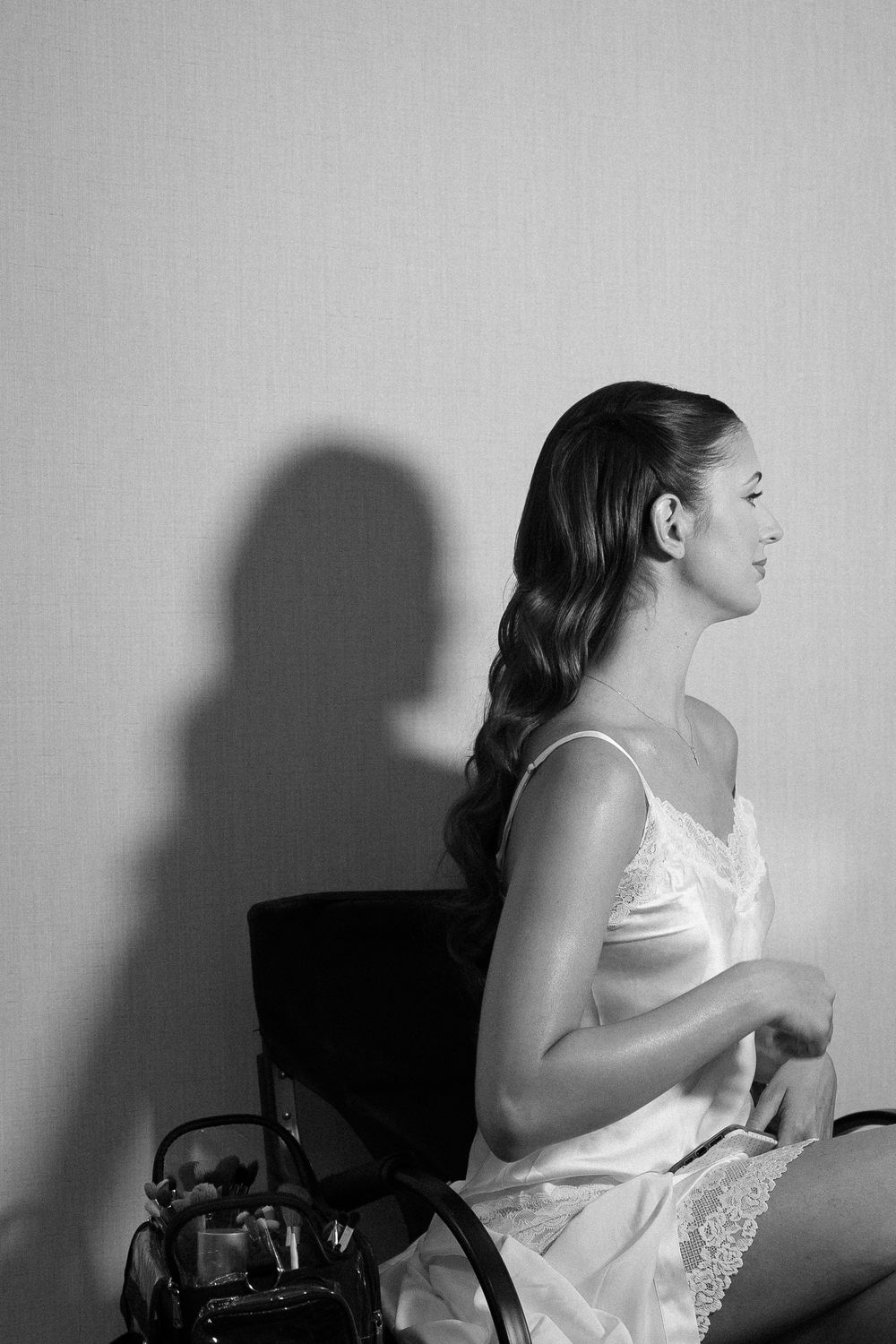 Black and white portrait of bride in white lace slip dress seated on chair with dramatic shadow on wall.
