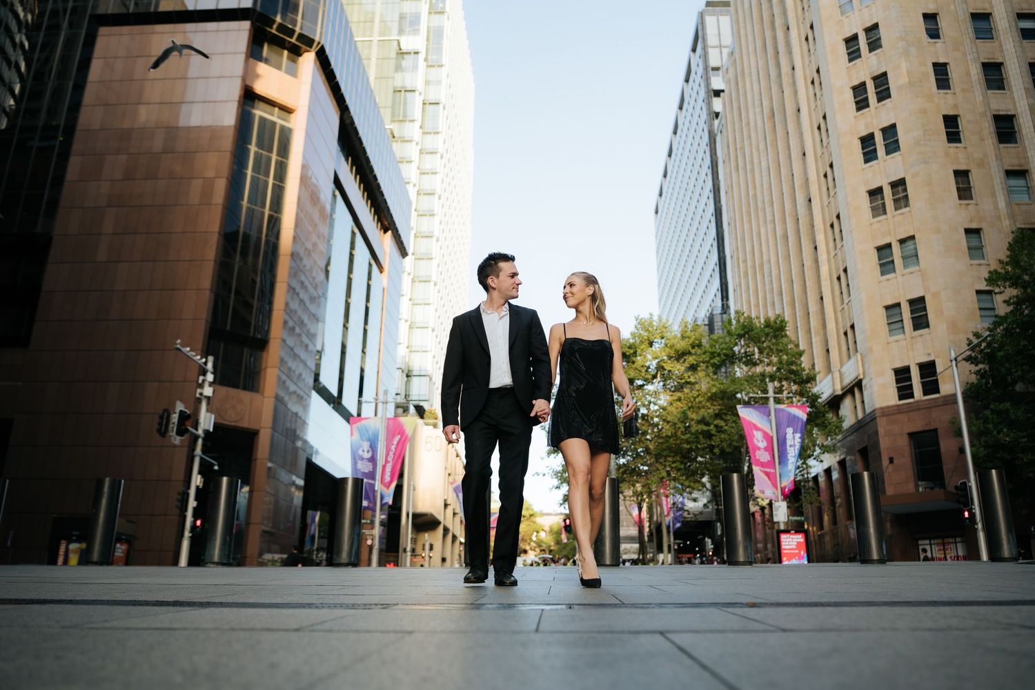 A well-dressed couple holds hands and walks along a modern city street lined with tall buildings.