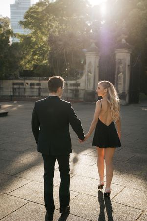 A couple in elegant black attire holds hands at sunset in front of ornate iron gates.