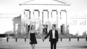 A couple holds hands walking in front of a grand neoclassical building in a black and white photo.