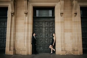 A stylish couple stands facing each other beside a grand bronze door of a sandstone building.