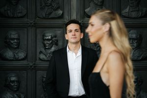 A man in a black suit gazes at a blonde woman standing before ornate bronze relief portrait panels.