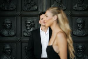 A man in a black suit looks thoughtfully past a blonde woman before decorative bronze portrait reliefs.