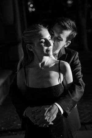 A man in a suit tenderly embraces and kisses a woman in a black dress in a dramatic black and white photo.