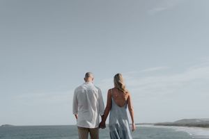 Couple holding hands facing the ocean on a serene beach with a pale blue sky.