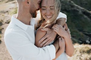 Happy couple embracing outdoors, woman smiling with engagement ring visible on her hand.