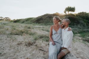 Couple sitting together on sand dunes at golden hour with grassy hills behind them.