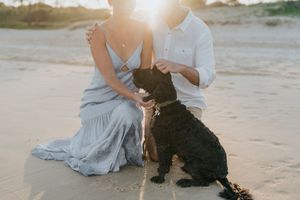 Couple sitting on beach sand with black curly dog at golden sunset.