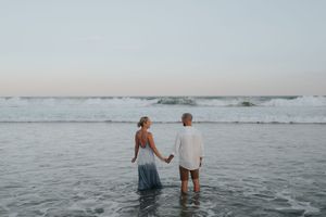 Couple holding hands standing in ocean waves at dusk on beach.
