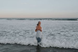 Woman standing in crashing ocean waves at dusk wearing blue dress.