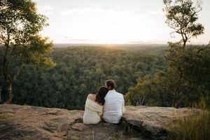 Couple sitting together on a rocky cliff edge watching a beautiful sunset over green forest.