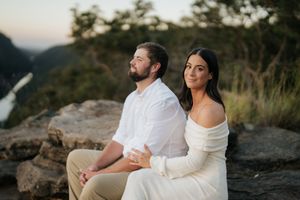 Couple sitting back to back on rocks with mountain river views during engagement photography.