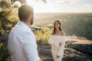 Woman in white off-shoulder dress smiling at partner during sunset engagement shoot outdoors.