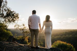 Couple holding hands from behind watching a golden sunset over a forested valley landscape.