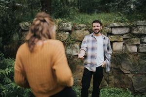 A smiling man in a plaid shirt reaches out his hand to a woman in an orange sweater in a lush garden setting.