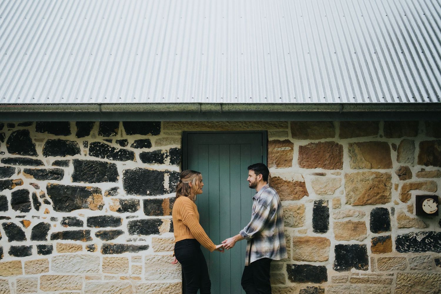 Couple holding hands facing each other beside a rustic stone building with green door.