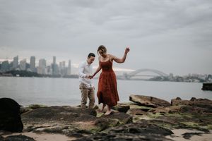 Couple playfully jumping on rocks by Sydney Harbour with the iconic bridge in the background.