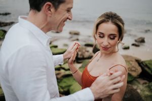 Couple sharing an intimate moment on rocky shoreline with moss-covered rocks and ocean behind.