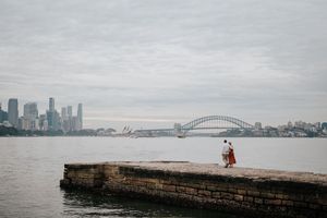 Couple stands on a stone pier with the iconic Sydney Harbour Bridge and Opera House in the background.