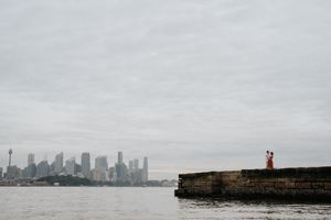 Couple standing on a stone pier with Sydney city skyline in the background on a cloudy day.