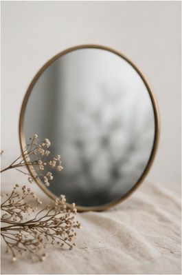 A round brass-framed mirror reflecting dried flowers, placed beside delicate baby's breath on linen fabric.