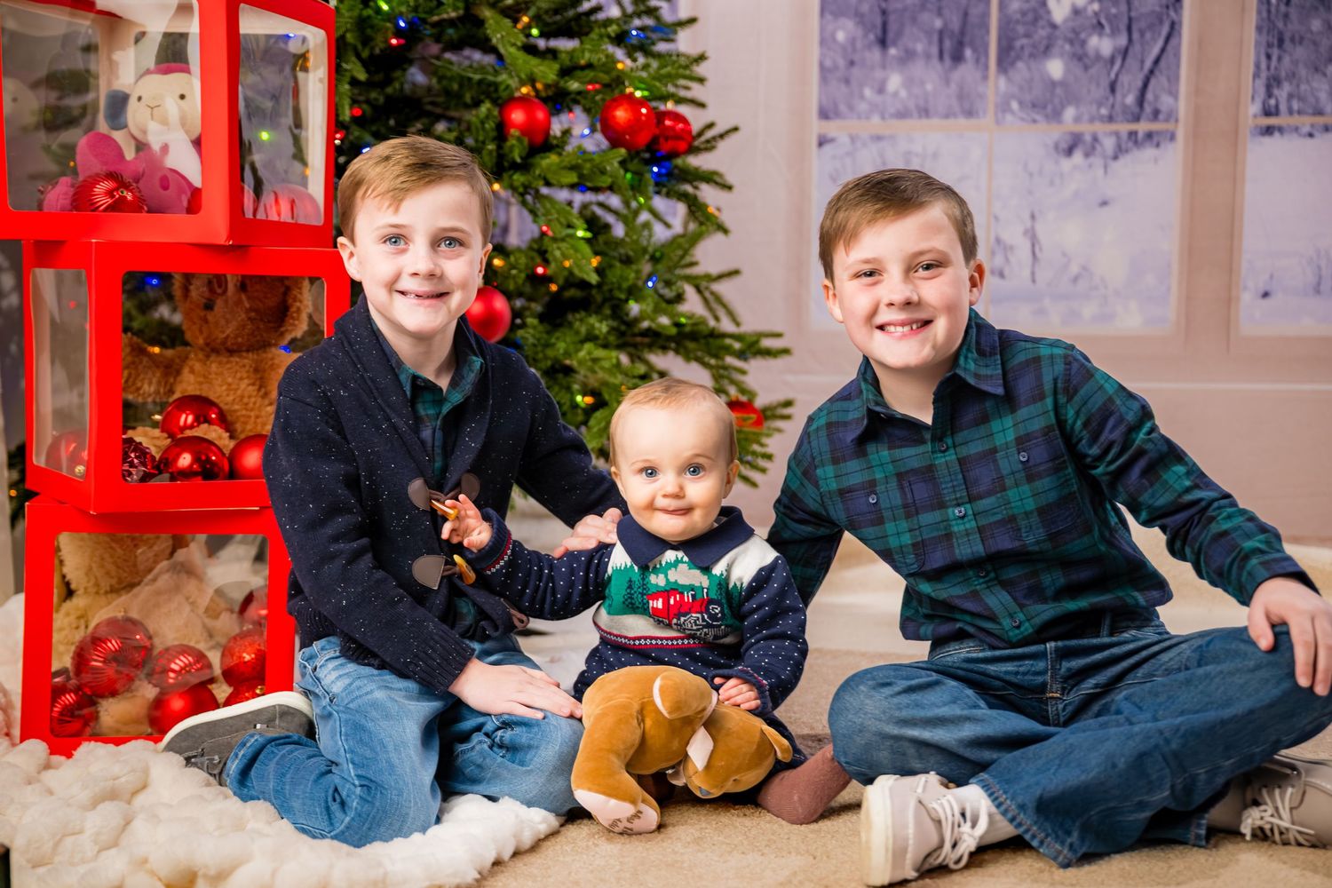 Three young boys pose for a Christmas photo with a decorated tree and holiday decorations in the background.
