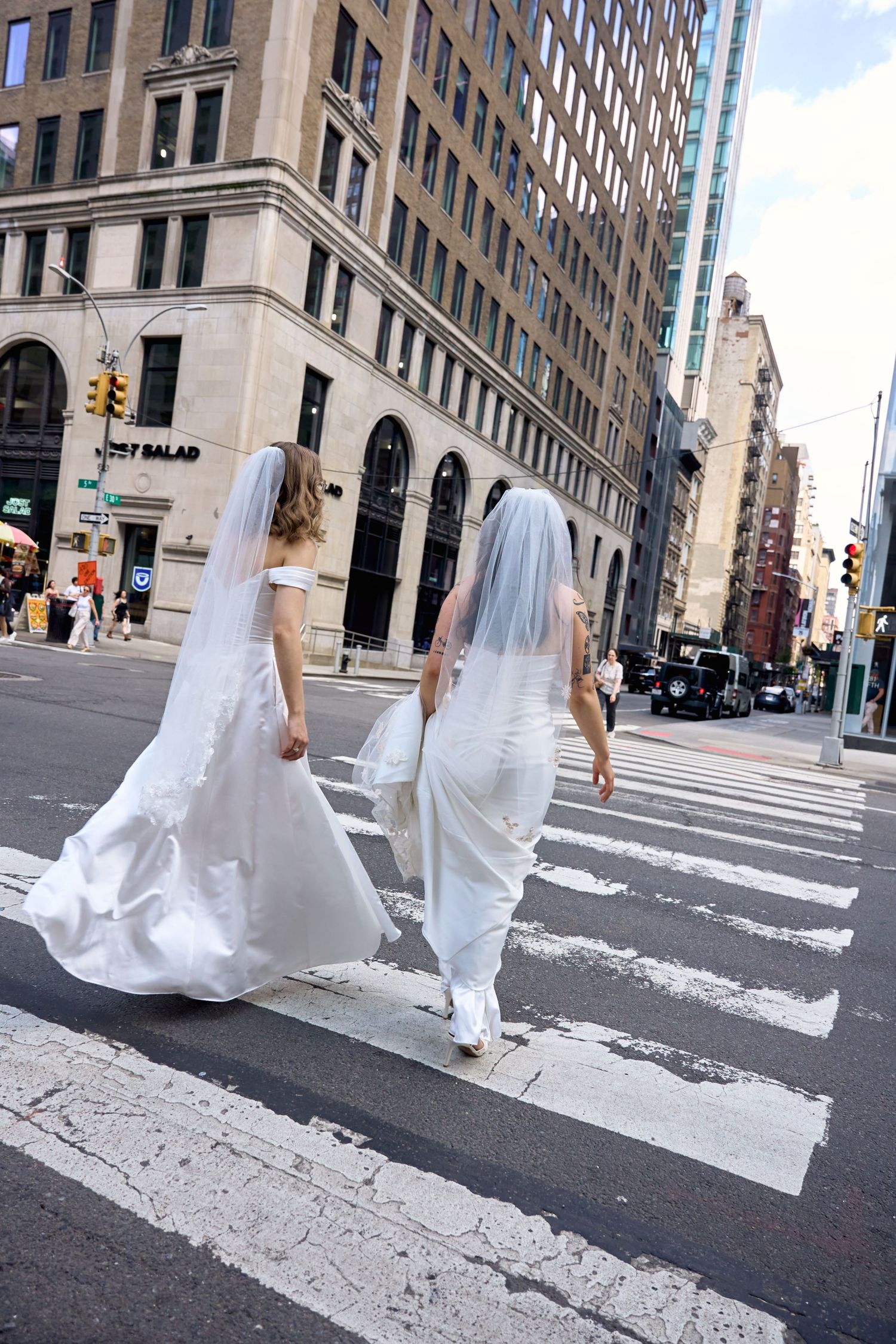 Two brides in white wedding gowns and veils walk together down a sunny city sidewalk.