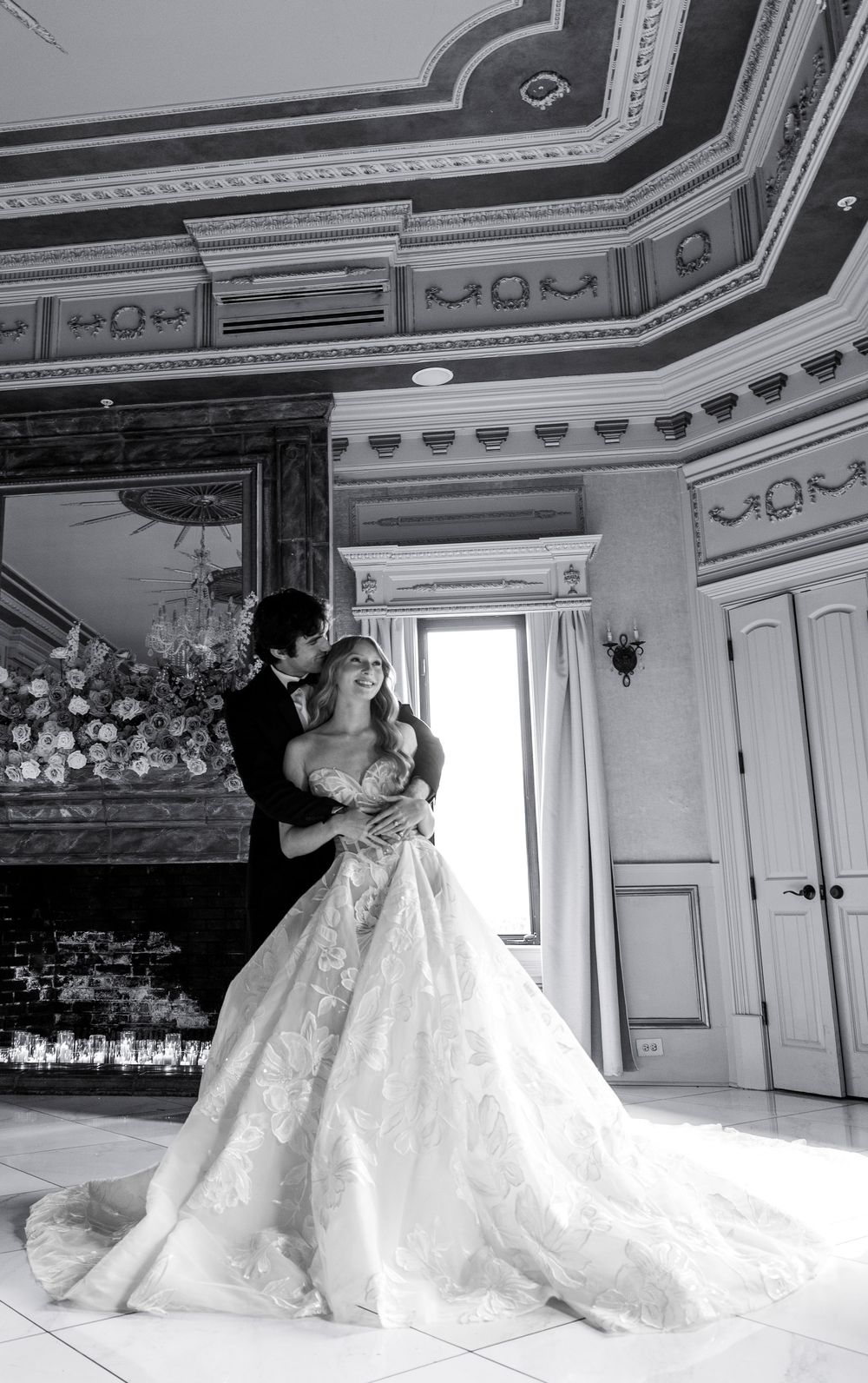 Black and white photo of bride and groom embracing in ornate ballroom with fireplace.