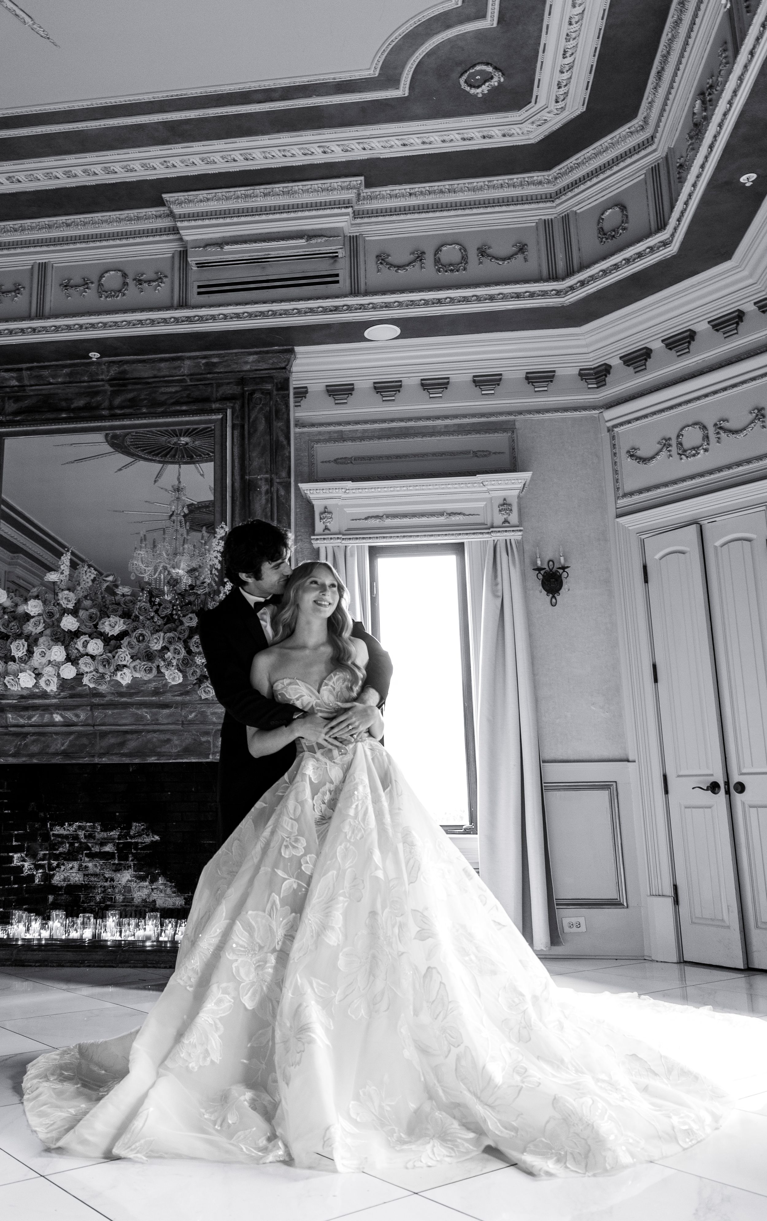 Black and white photo of bride and groom embracing in ornate ballroom with fireplace.