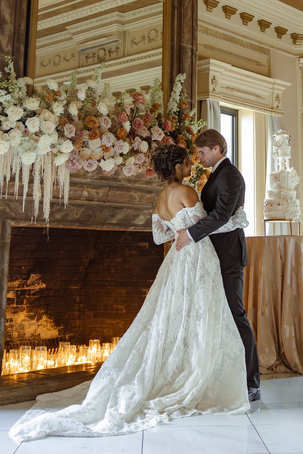 Bride and groom embrace before floral fireplace with candles in elegant wedding venue.