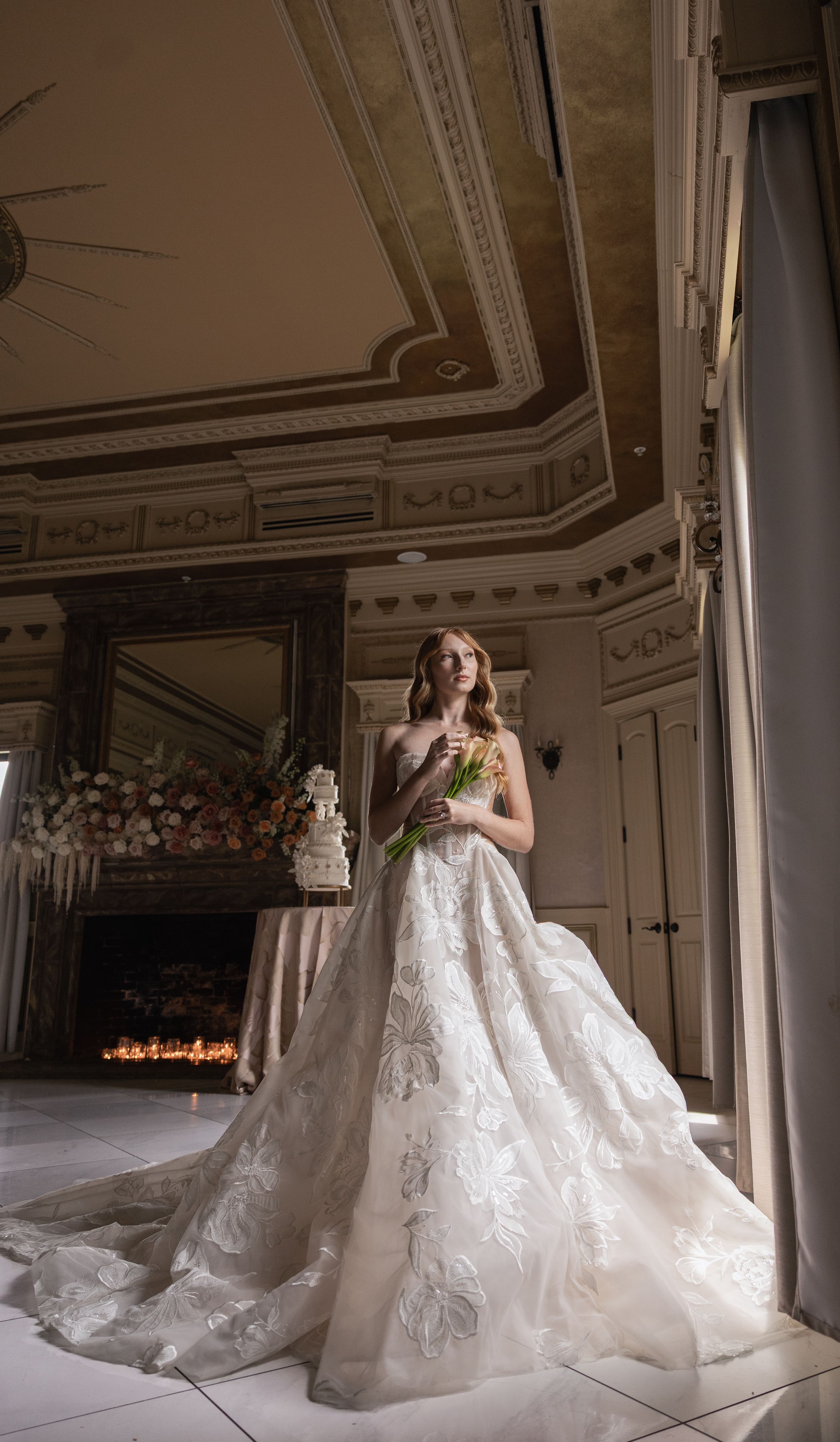 Bride in ornate lace ball gown holding bouquet in elegant venue with decorative ceiling and fireplace.